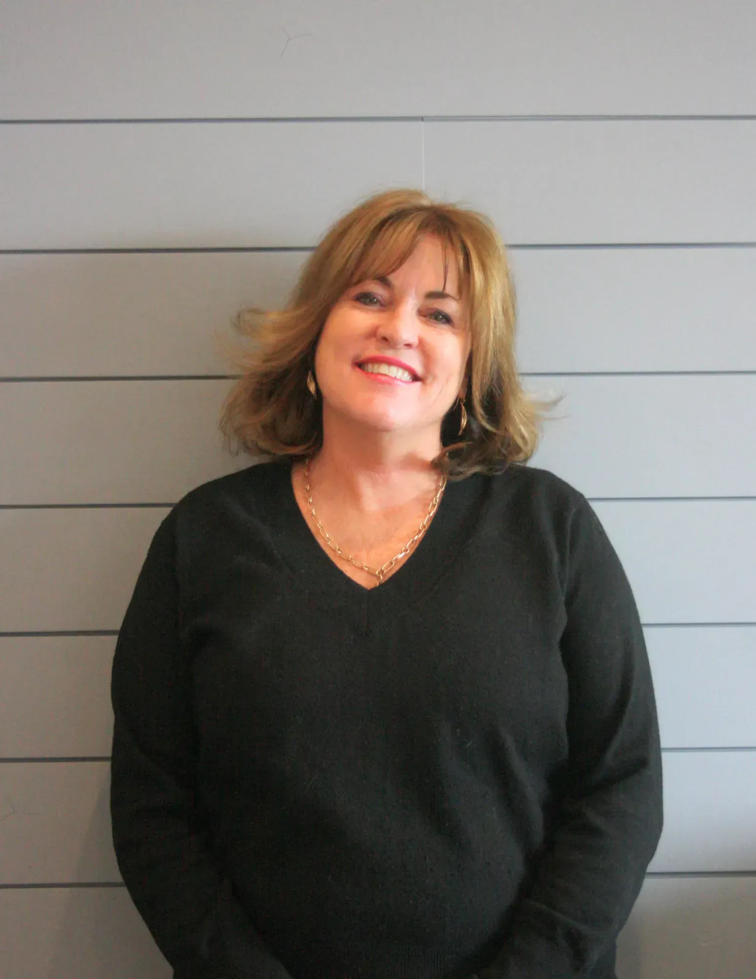 Smiling person in a black sweater standing against a gray wall.