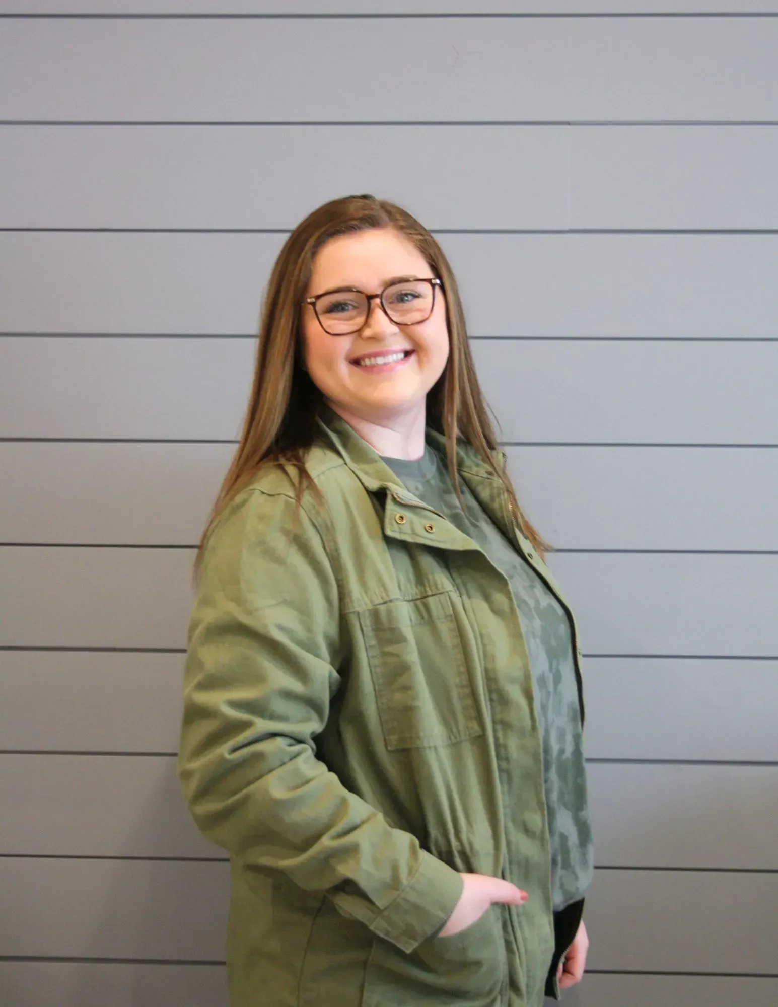 Smiling person in glasses wearing a green jacket, standing against a gray wall.
