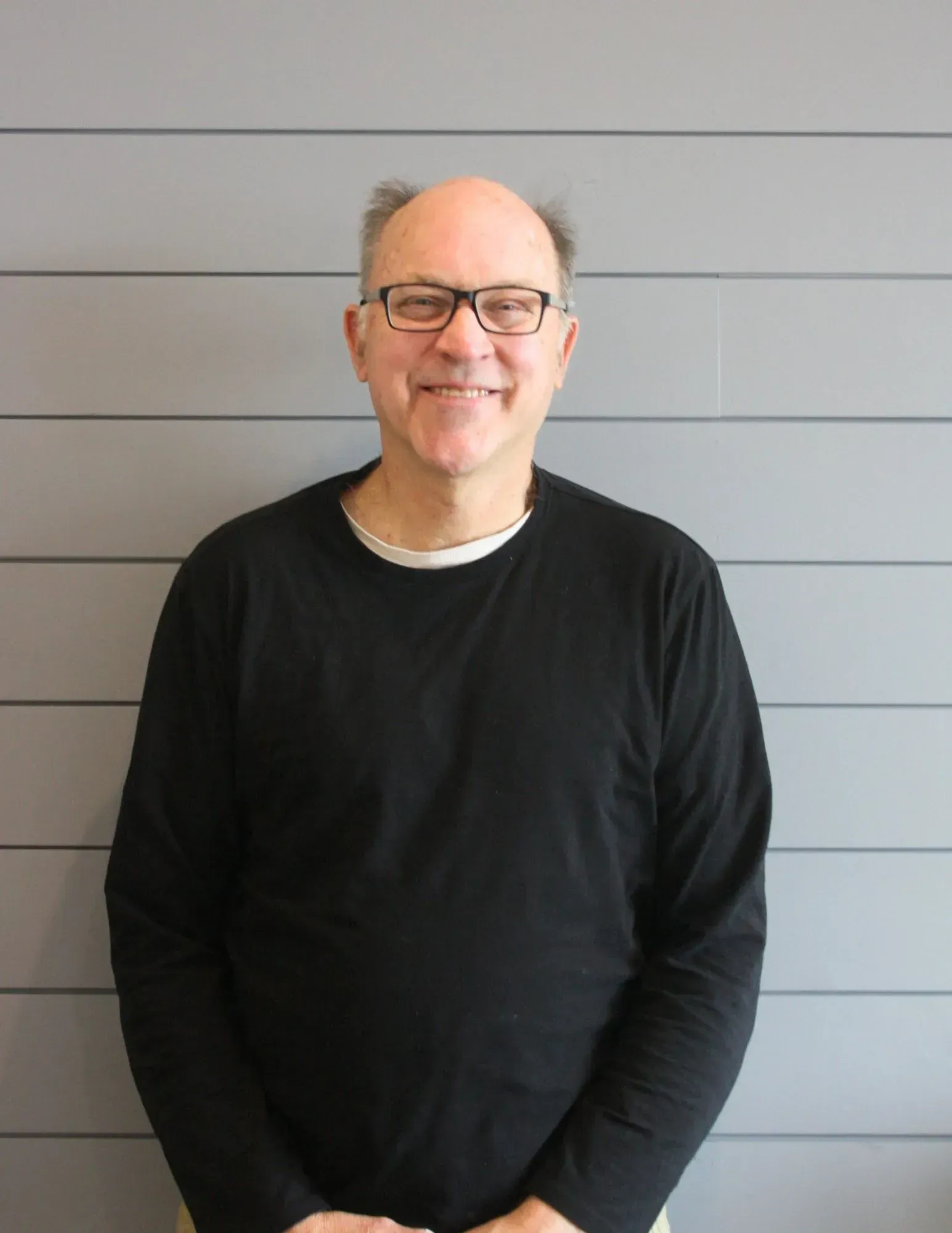 Smiling person in glasses wearing a black shirt, standing against a gray paneled wall.
