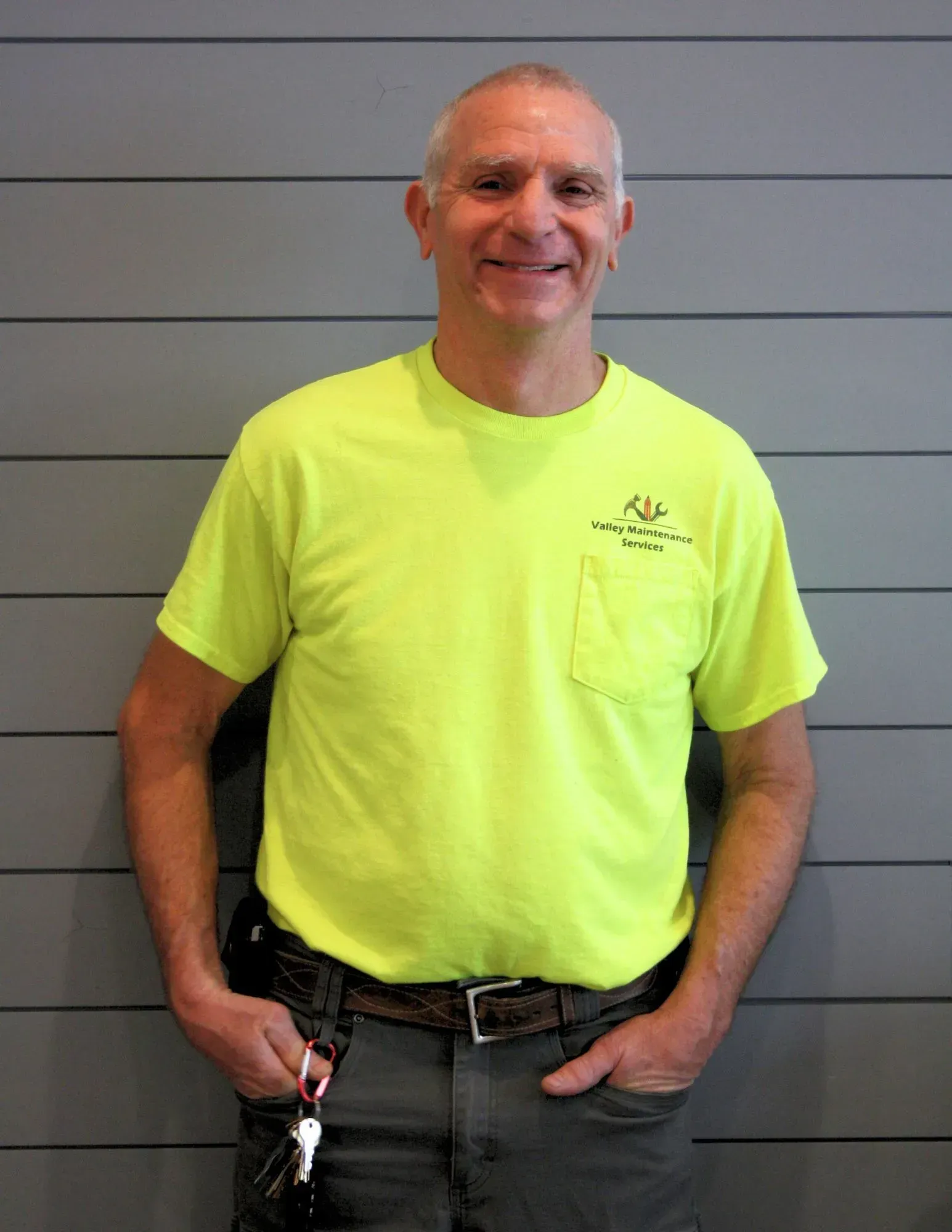 Smiling person in a neon yellow T-shirt and gray pants standing against a gray wall.