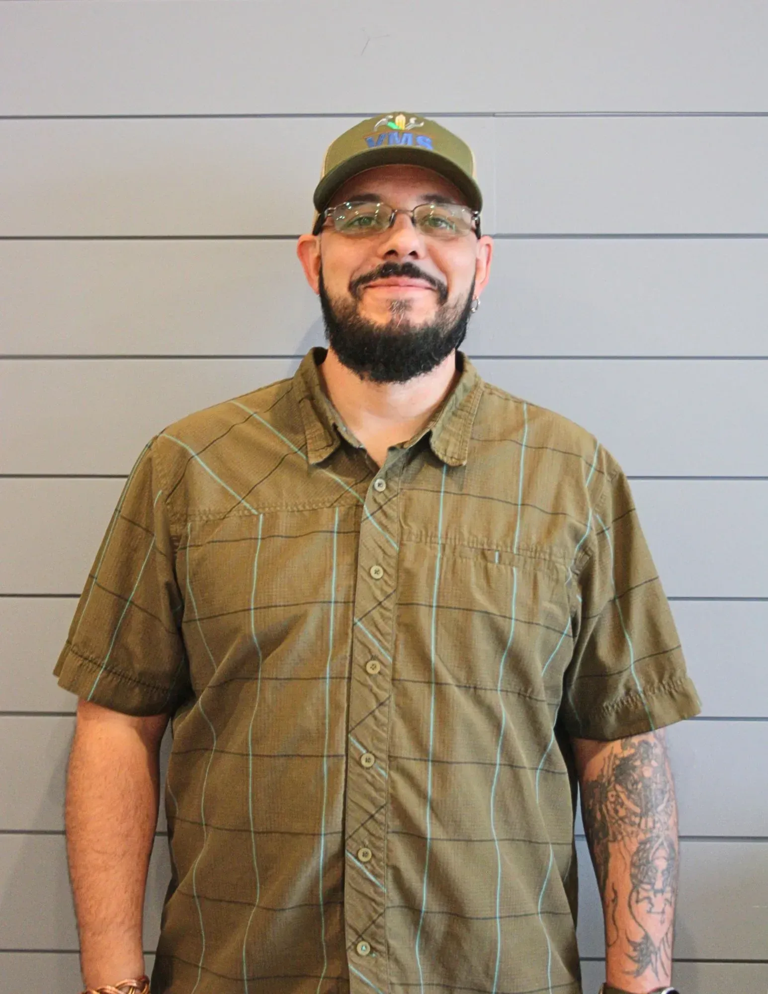Man standing against a gray wall, wearing a green plaid shirt and cap, with a tattooed right arm.