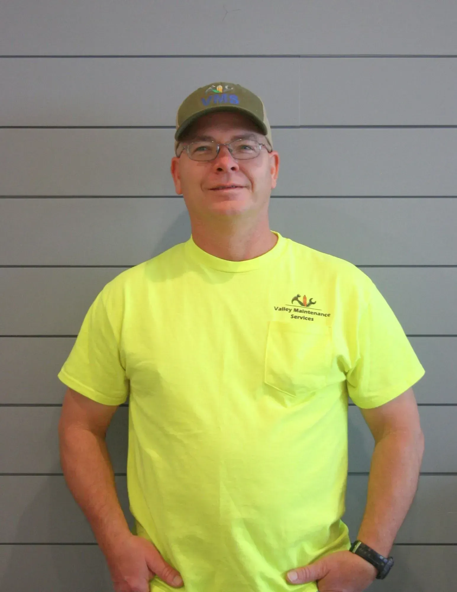 Person in neon yellow T-shirt and cap standing against a gray wall, hands in pockets.