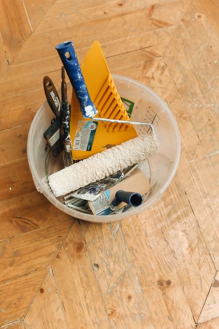 A bowl on a wooden floor holds assorted household tools, including a blue handle and yellow scrubber.