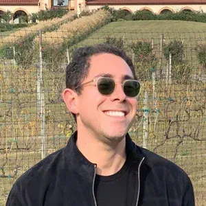 Smiling person in sunglasses and a black jacket standing outdoors in a vineyard