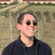 Smiling person in sunglasses and a black jacket standing outdoors in a vineyard