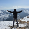 Person standing on a snowy mountain with arms outstretched, overlooking distant peaks under a blue sky