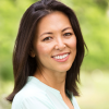Smiling woman with shoulder-length dark hair in a light top, standing outdoors against a blurred green background