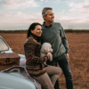 Two people holding a small dog beside a car in a rural field at sunset