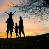 Silhouetted family standing on a hill at sunset beside a tree, with glowing orange sky and clouds.