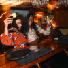 Two people in orange jackets smiling at a bar with drinks and warm hanging lights