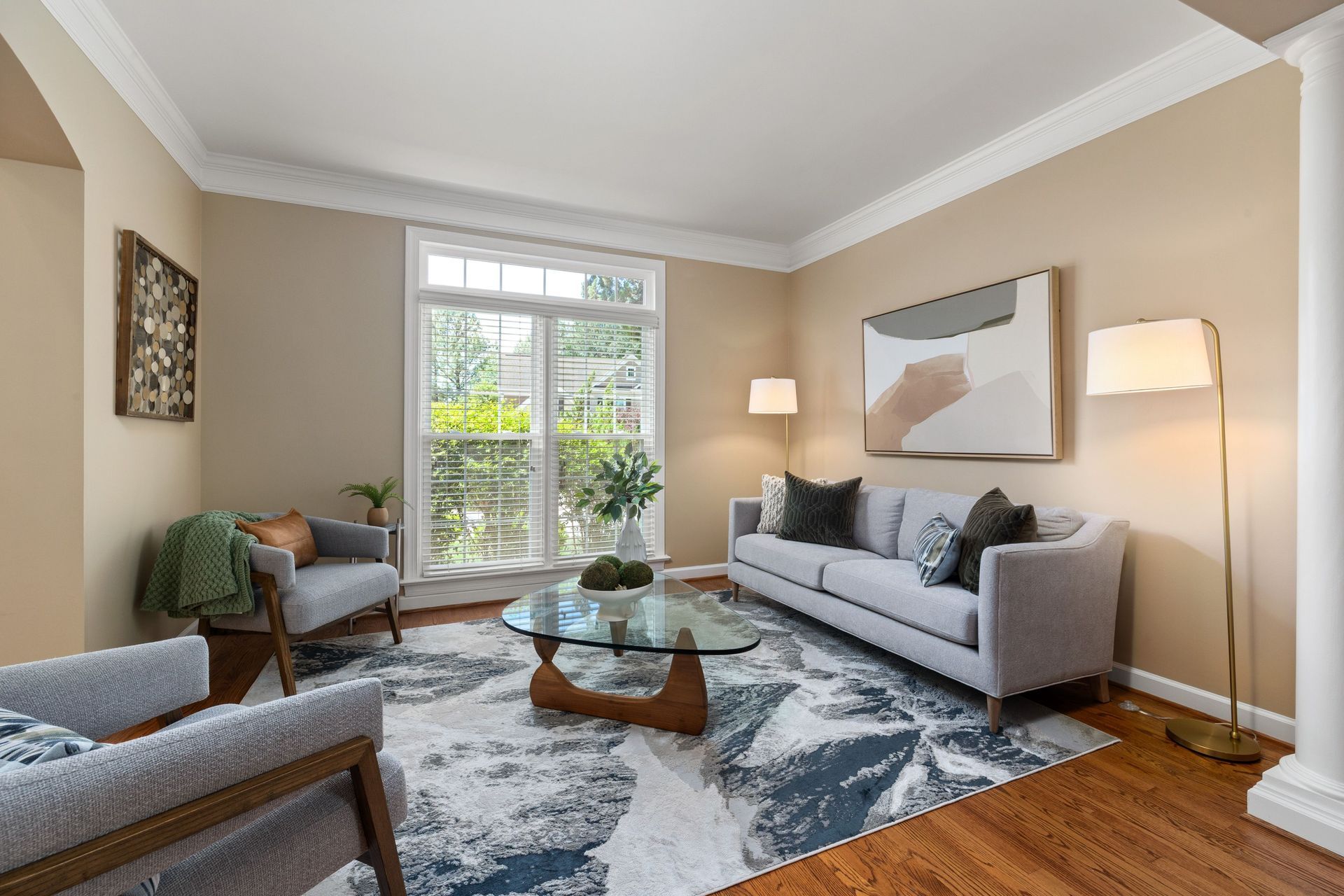 Bright living room with gray sofa, armchair, glass coffee table, and large window with greenery view.