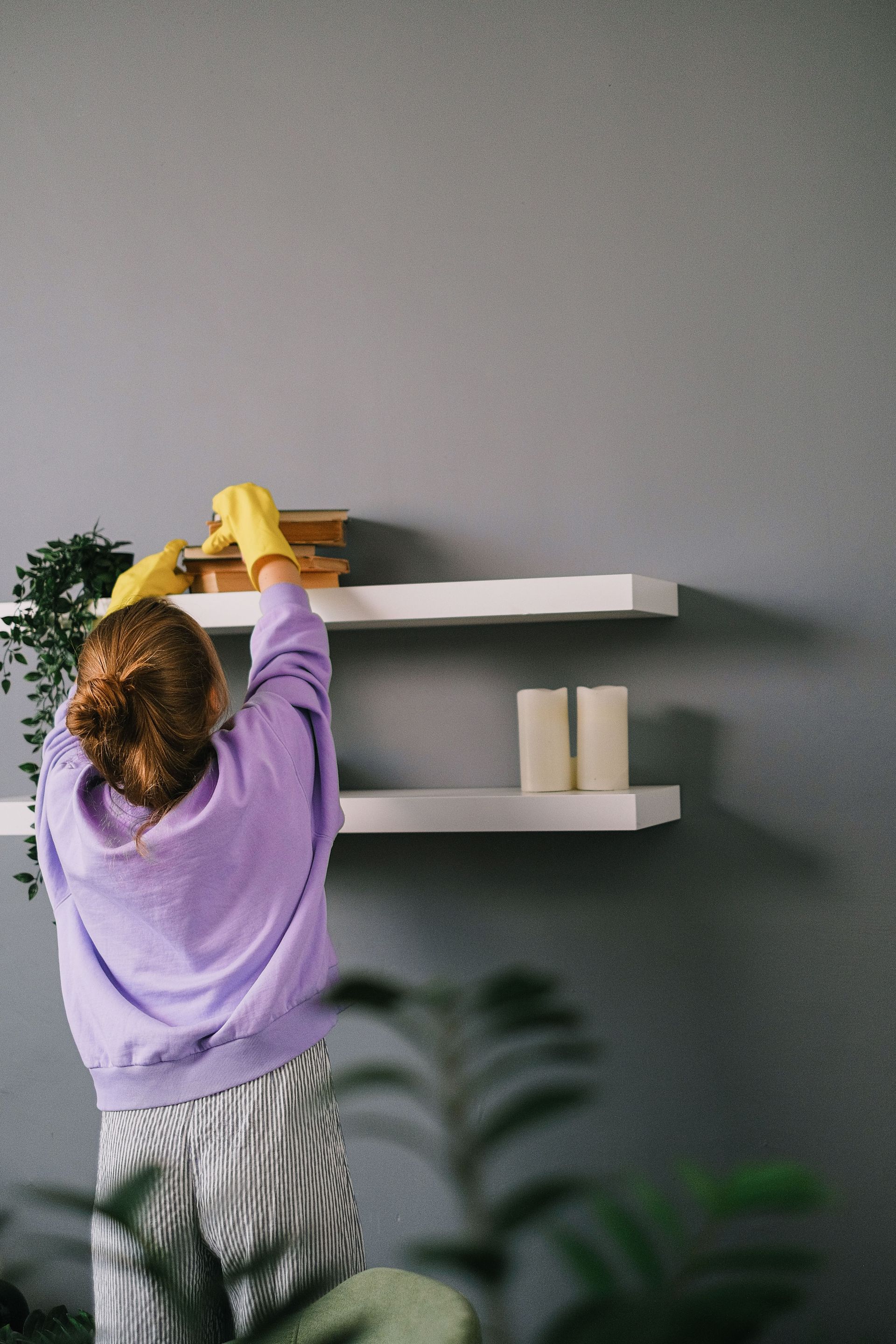 Person in a purple sweater cleaning a white wall shelf in a gray room with a yellow cloth