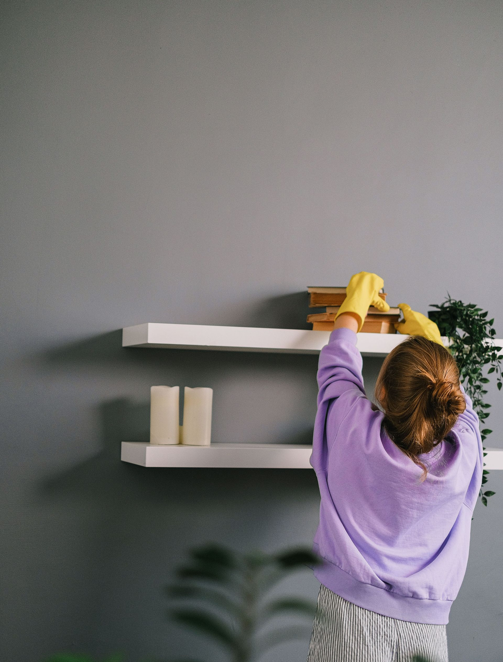 Person in a purple sweater dusts white wall shelves with a yellow cloth.