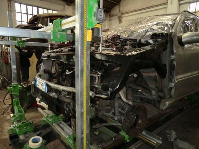 Carrozzeria scura e schiacciata in un'officina, sorretta da supporti verdi, con attrezzi nelle vicinanze.