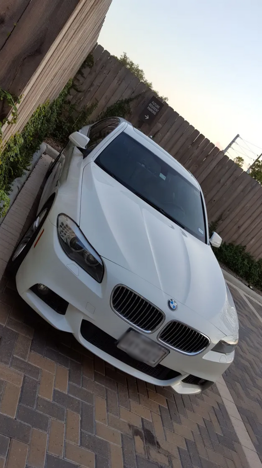 White BMW sedan parked on a brick driveway beside a wooden fence and garden wall.