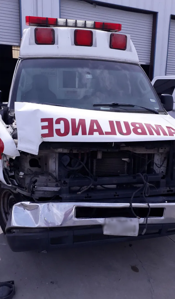 Damaged ambulance with crushed front hood and missing grille, parked on a street.