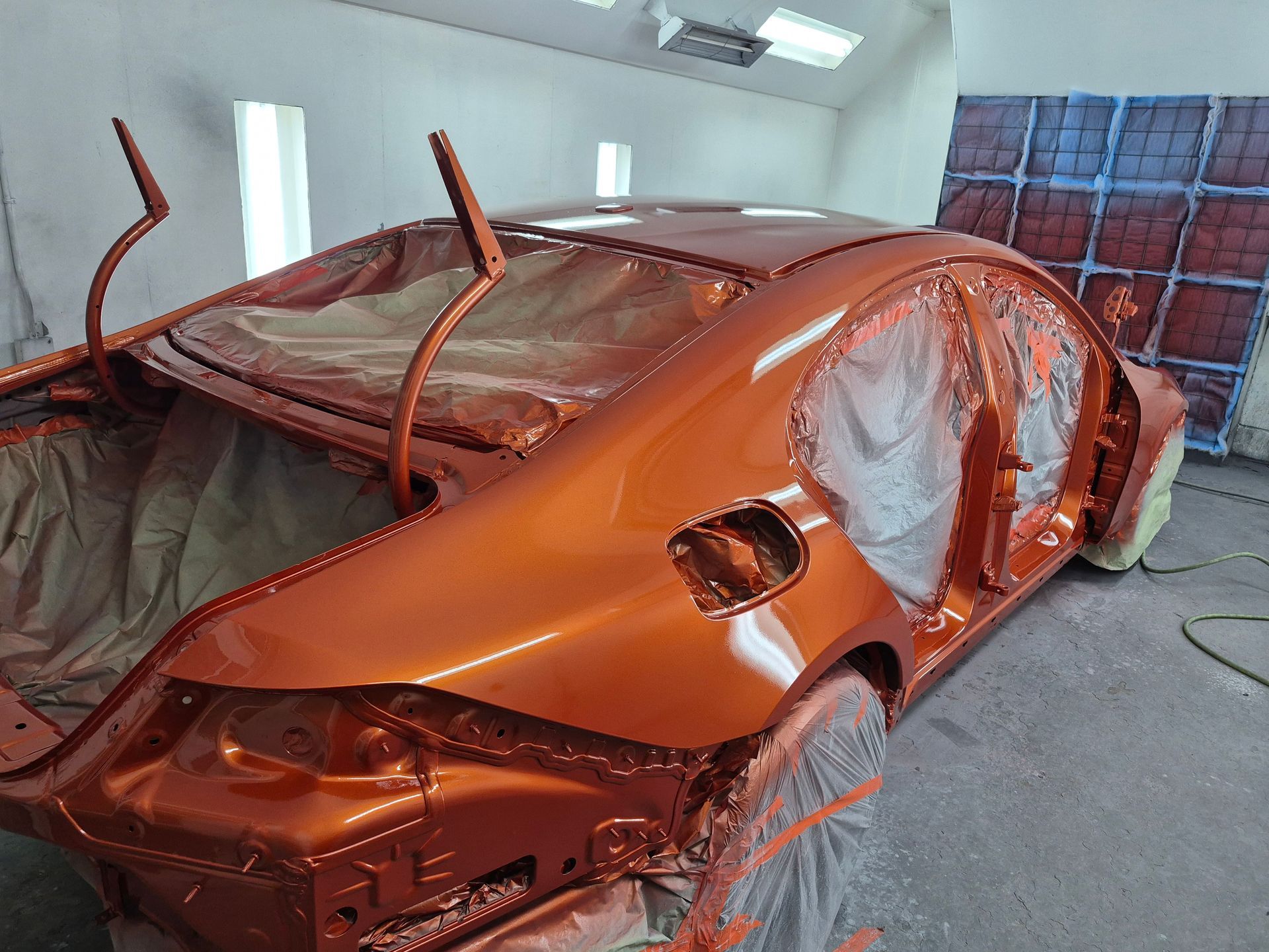Orange car body in a paint booth, covered with masking plastic and wet paint overspray visible