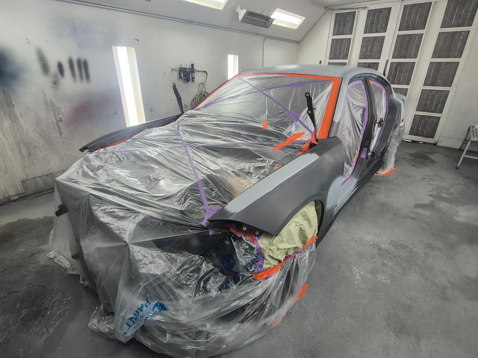 Car covered in plastic and tape inside an auto body paint booth, with red framing and a damaged front end