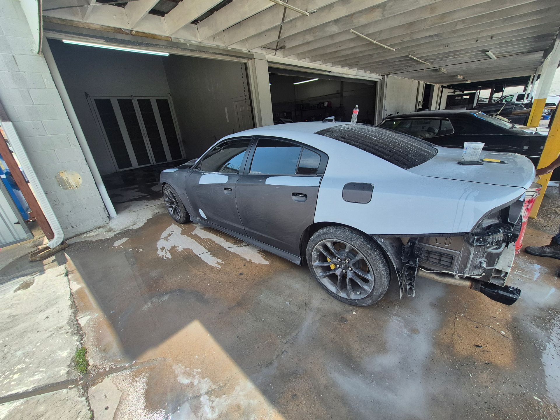 Grey sedan parked in a wet garage, heavily damaged with a detached rear bumper and open trunk area