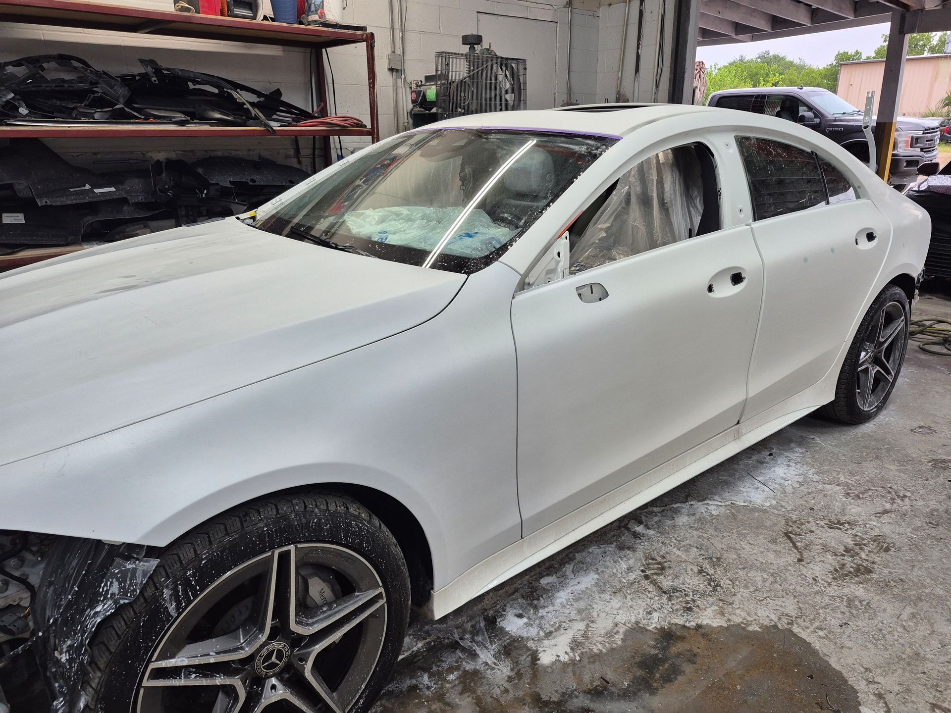 White sedan in a workshop, partially painted with black rear wheel and open garage equipment nearby