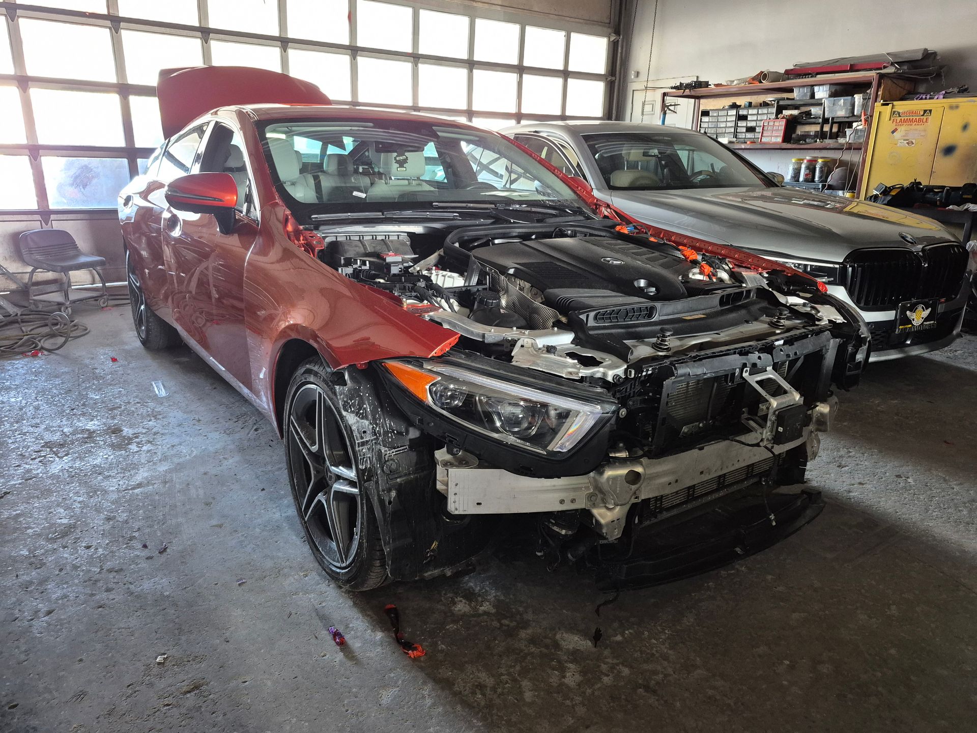 Damaged red car in an auto repair shop, front end disassembled with hood open.