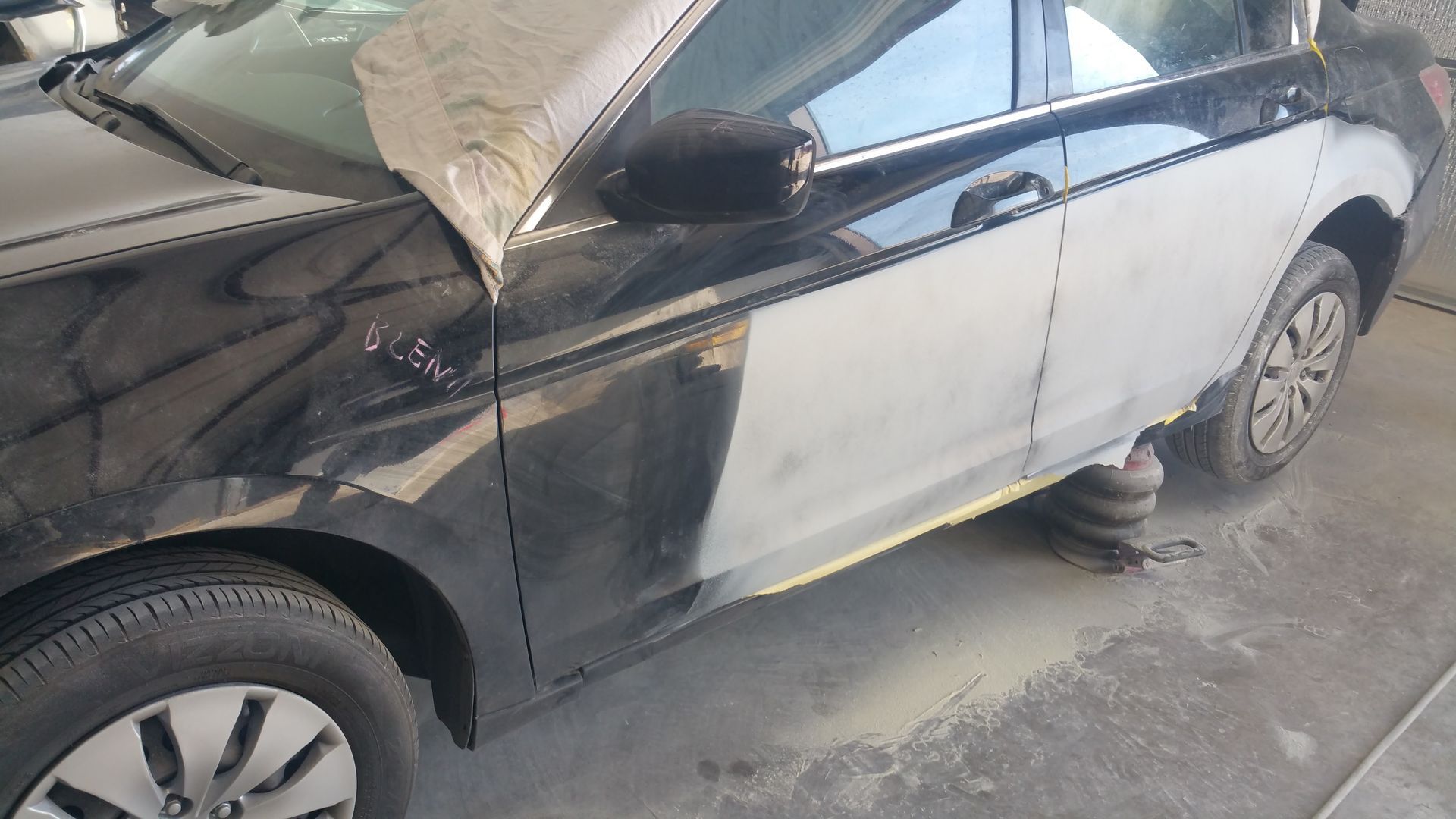 Black and white sedan with visible front-end and side damage, parked in a garage over a floor jack