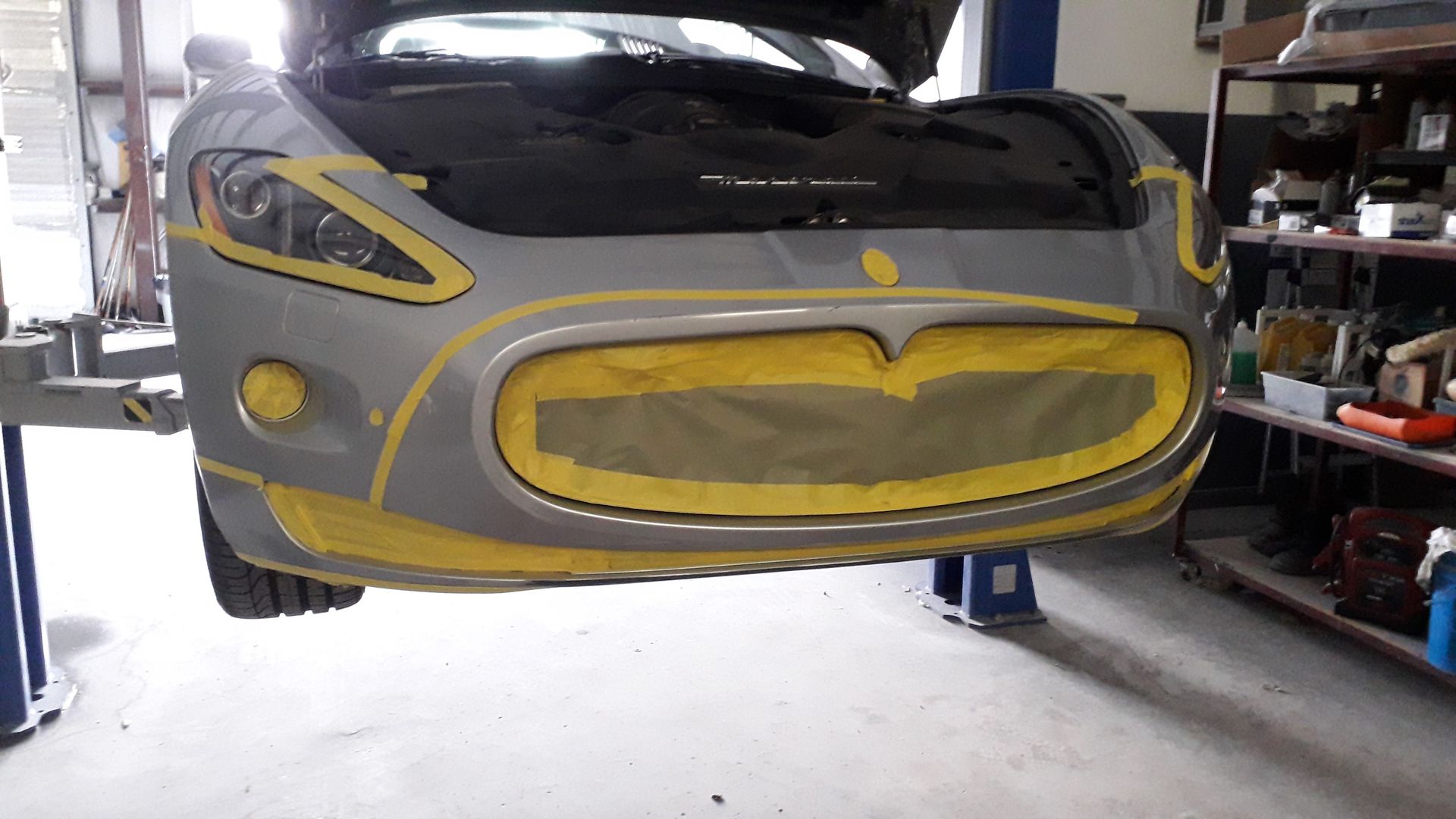Front of a gray car wrapped in yellow tape, lifted in a garage or workshop.