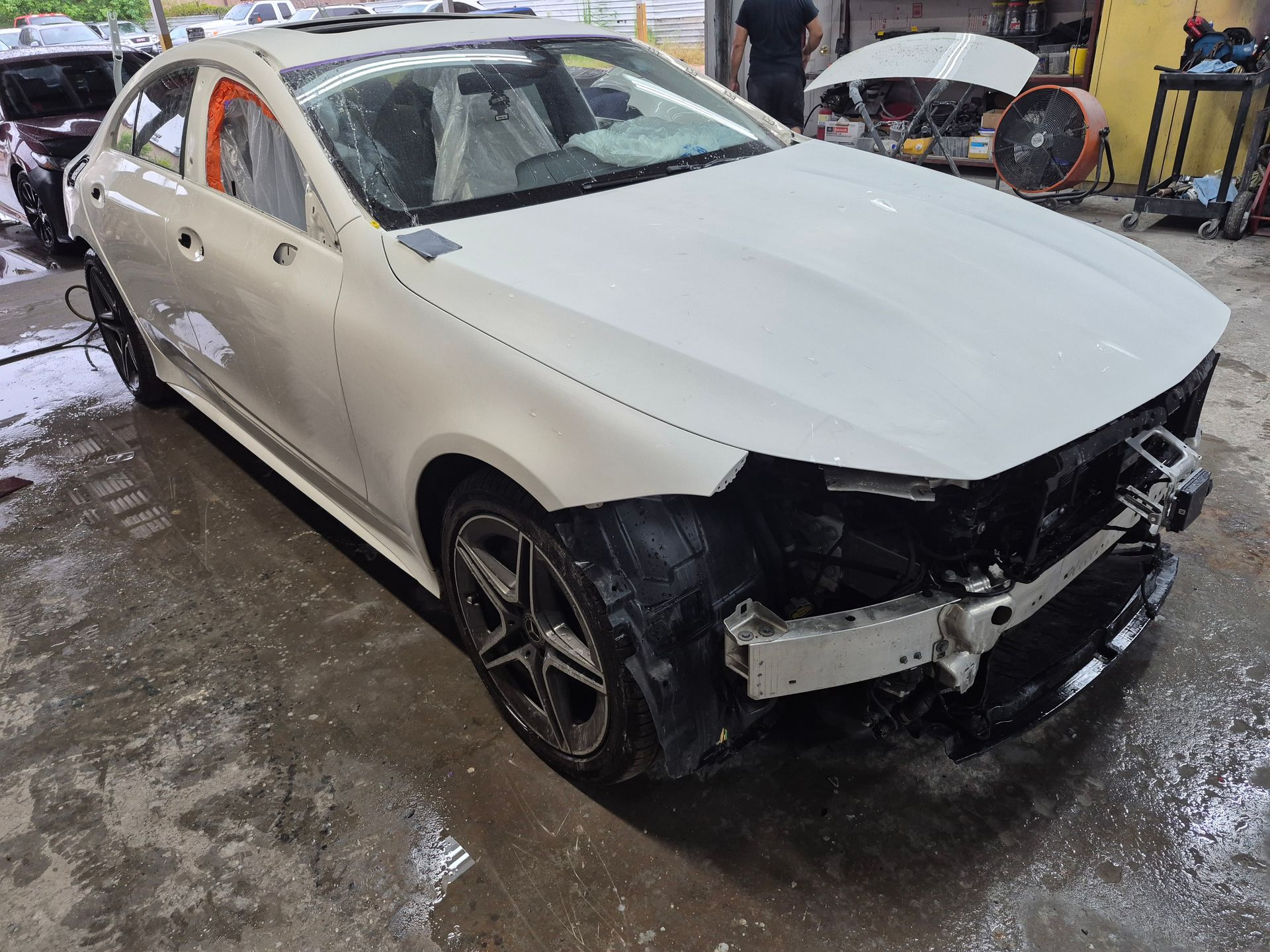 White sedan with front-end damage in a wet auto repair shop, hood open and bumper removed
