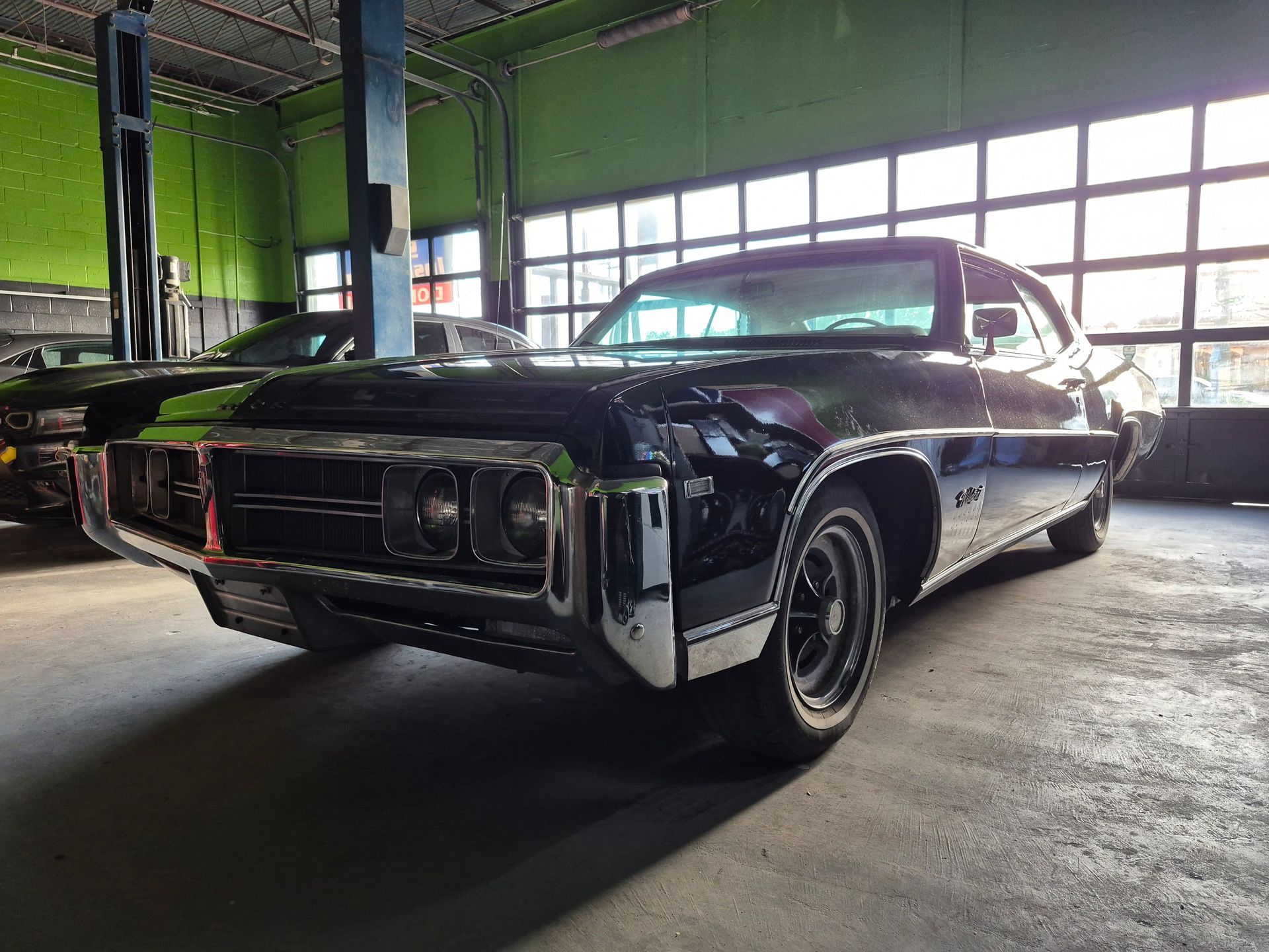 Classic black lowrider car in a garage, shown front-left under bright industrial windows.