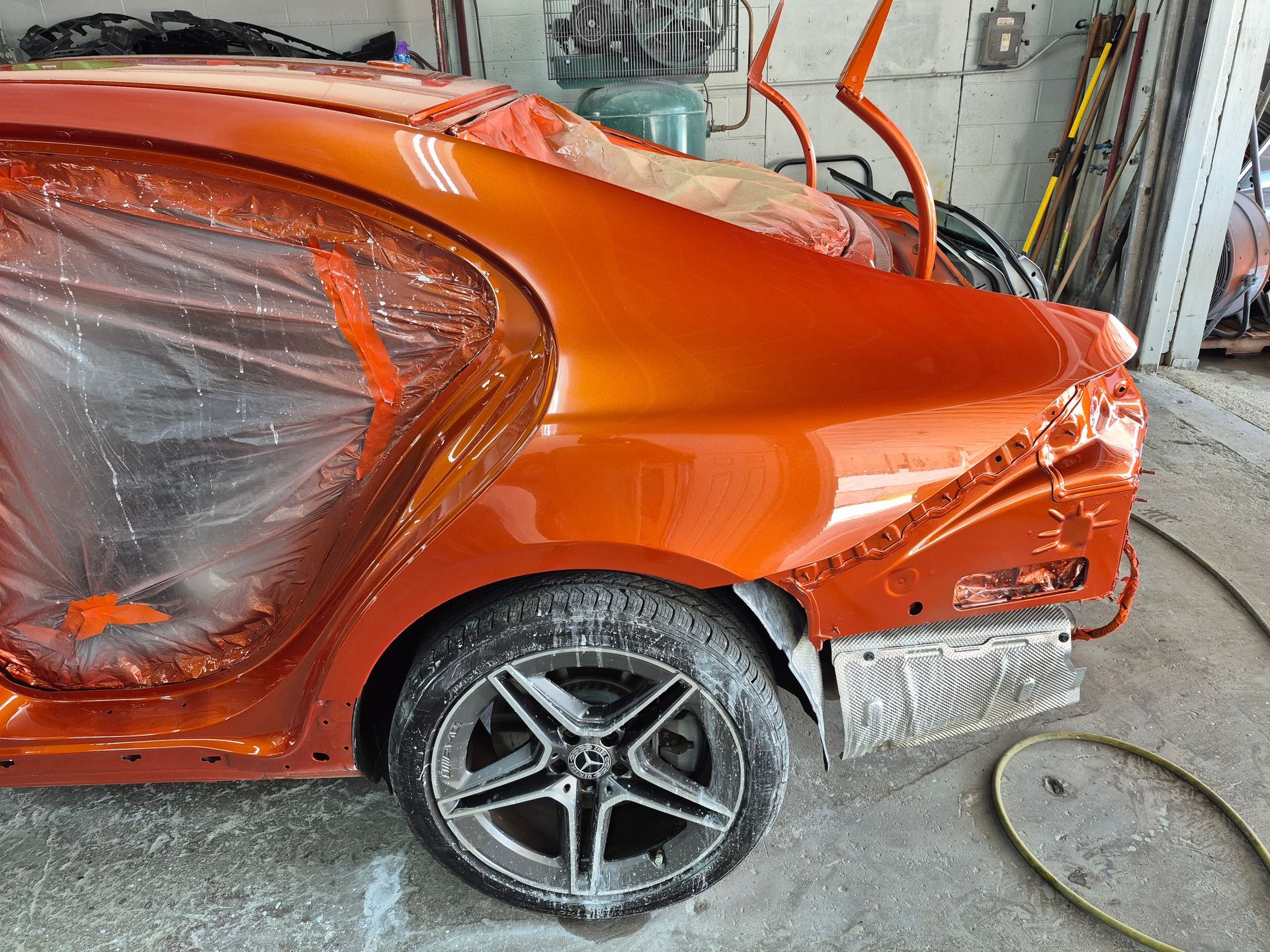 Orange car with rear damage in a repair shop, wheel exposed and covered in plastic