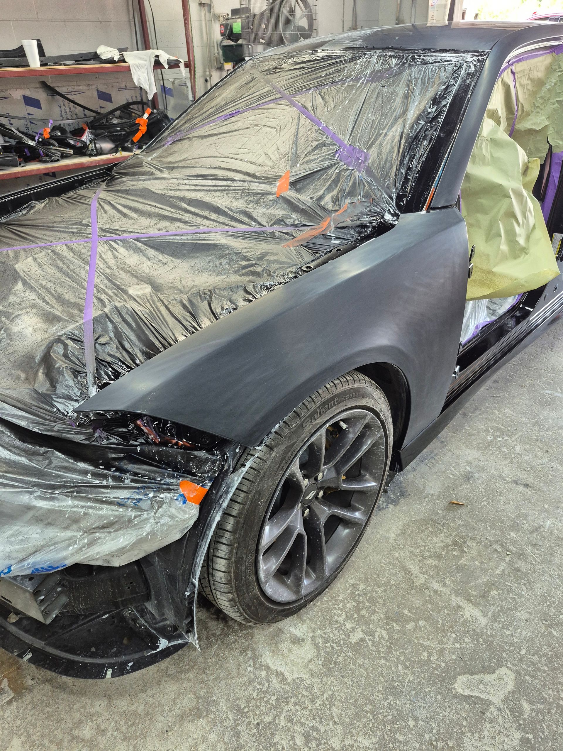 Black car with windshield wrapped in plastic and purple straps, parked in a garage or body shop after damage.