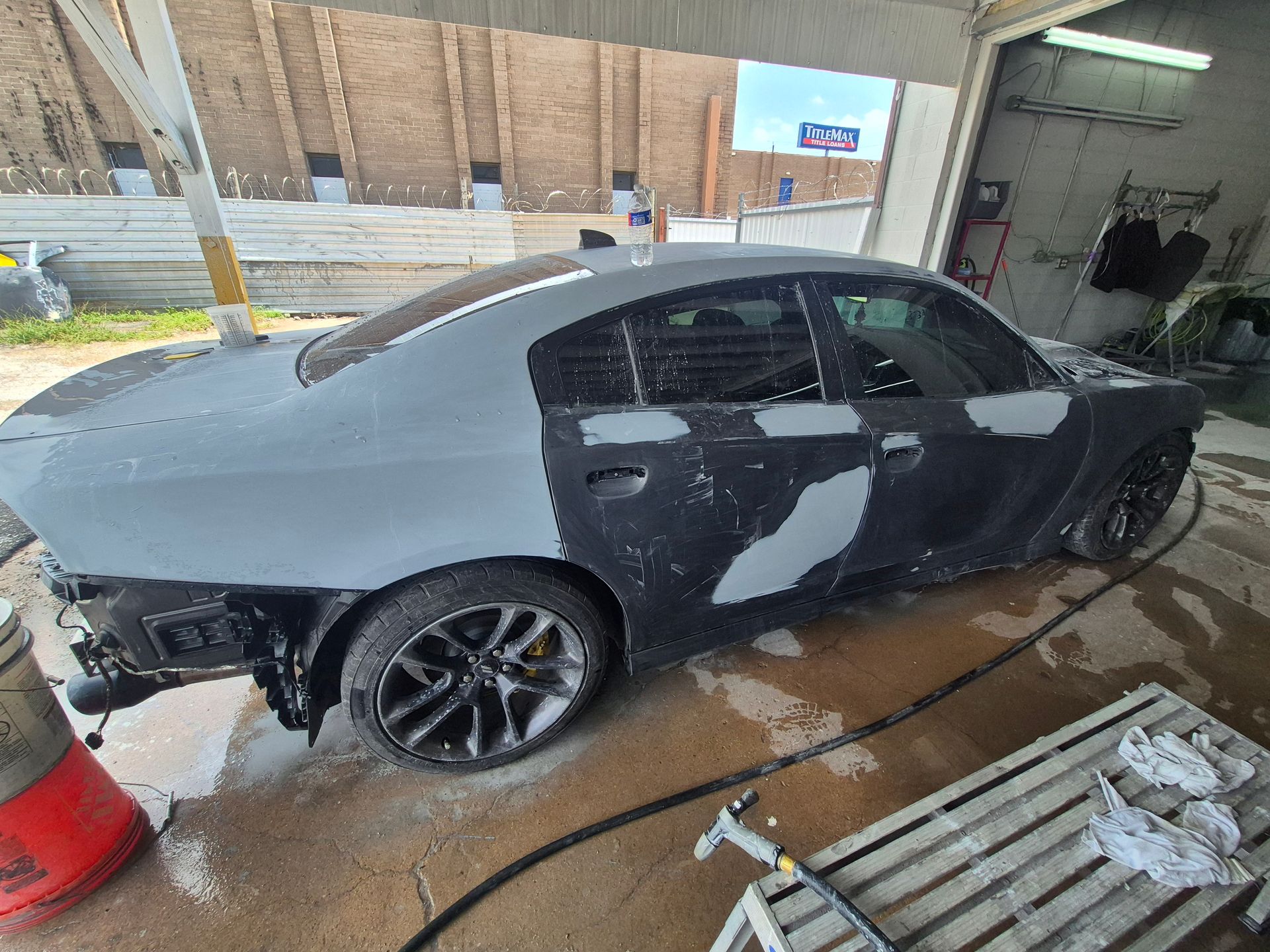 Black sedan in a car wash bay, covered in soap and water, with a hose and cleaning tools nearby.