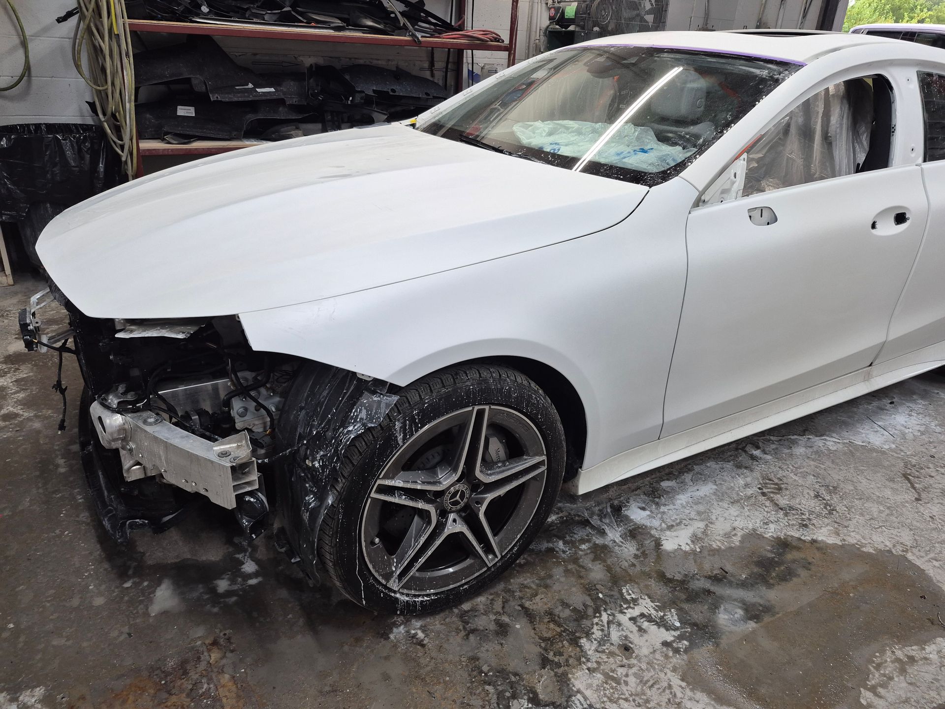 White sedan with front-left bumper damage parked in a wet auto repair shop bay
