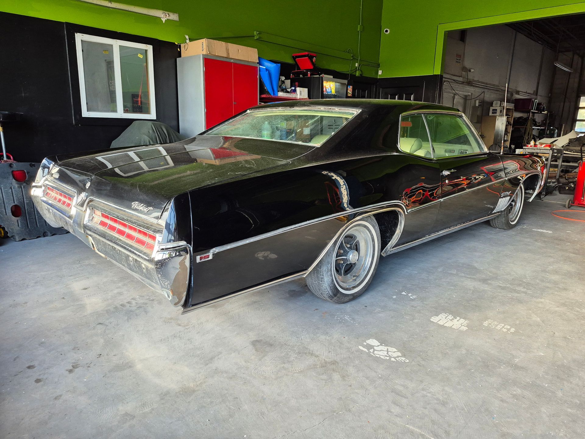 Black classic car in a garage, viewed from the rear side, with chrome trim and red interior visible