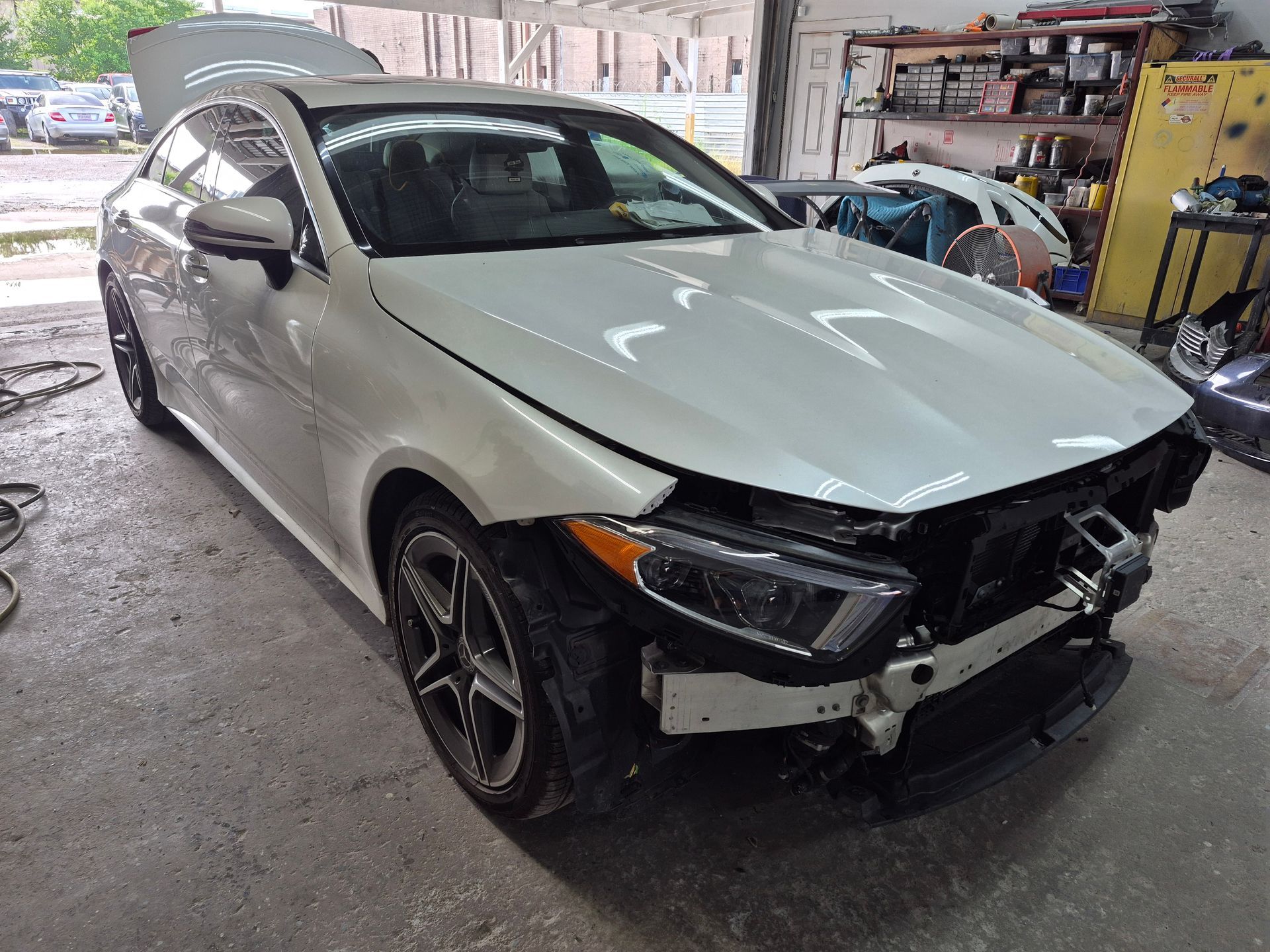 White sedan with front-end damage in an auto repair shop, missing bumper and grille