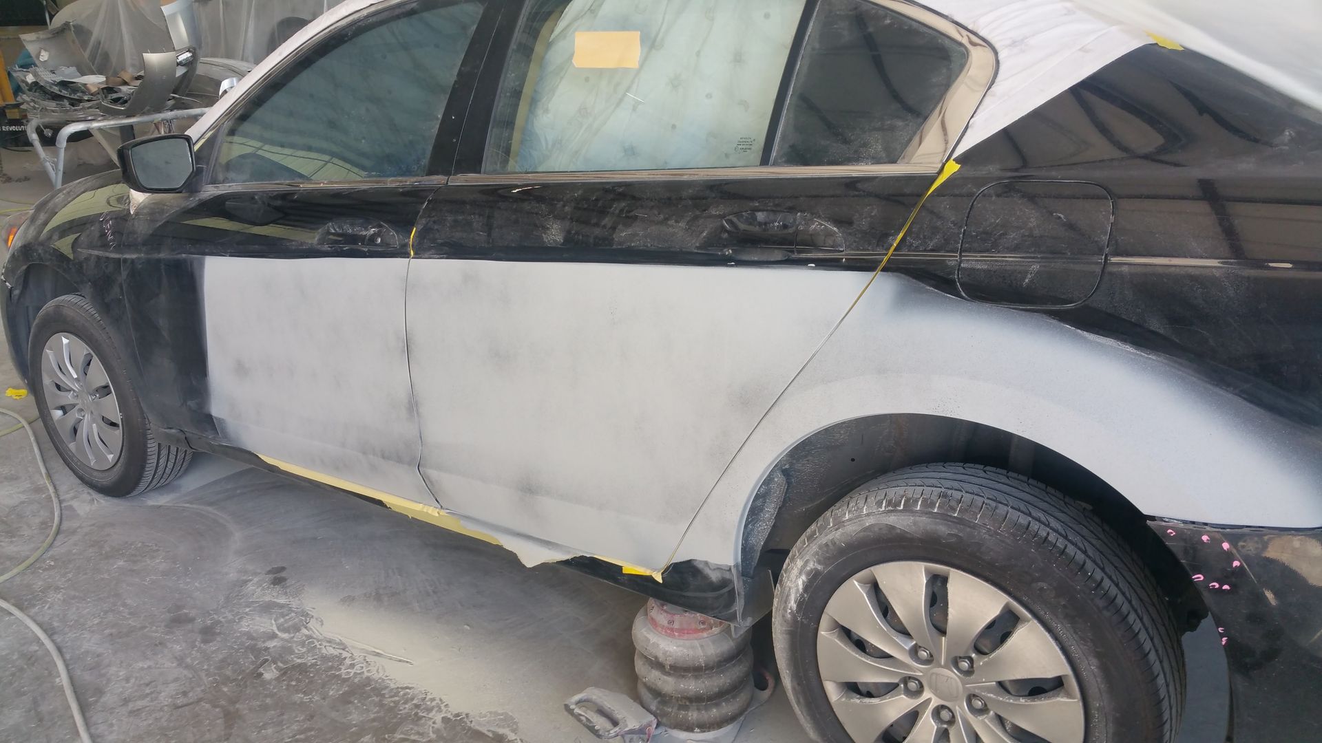 Black car with a gray primer-painted side panel in a repair shop, wheels visible and covered partly by tape.