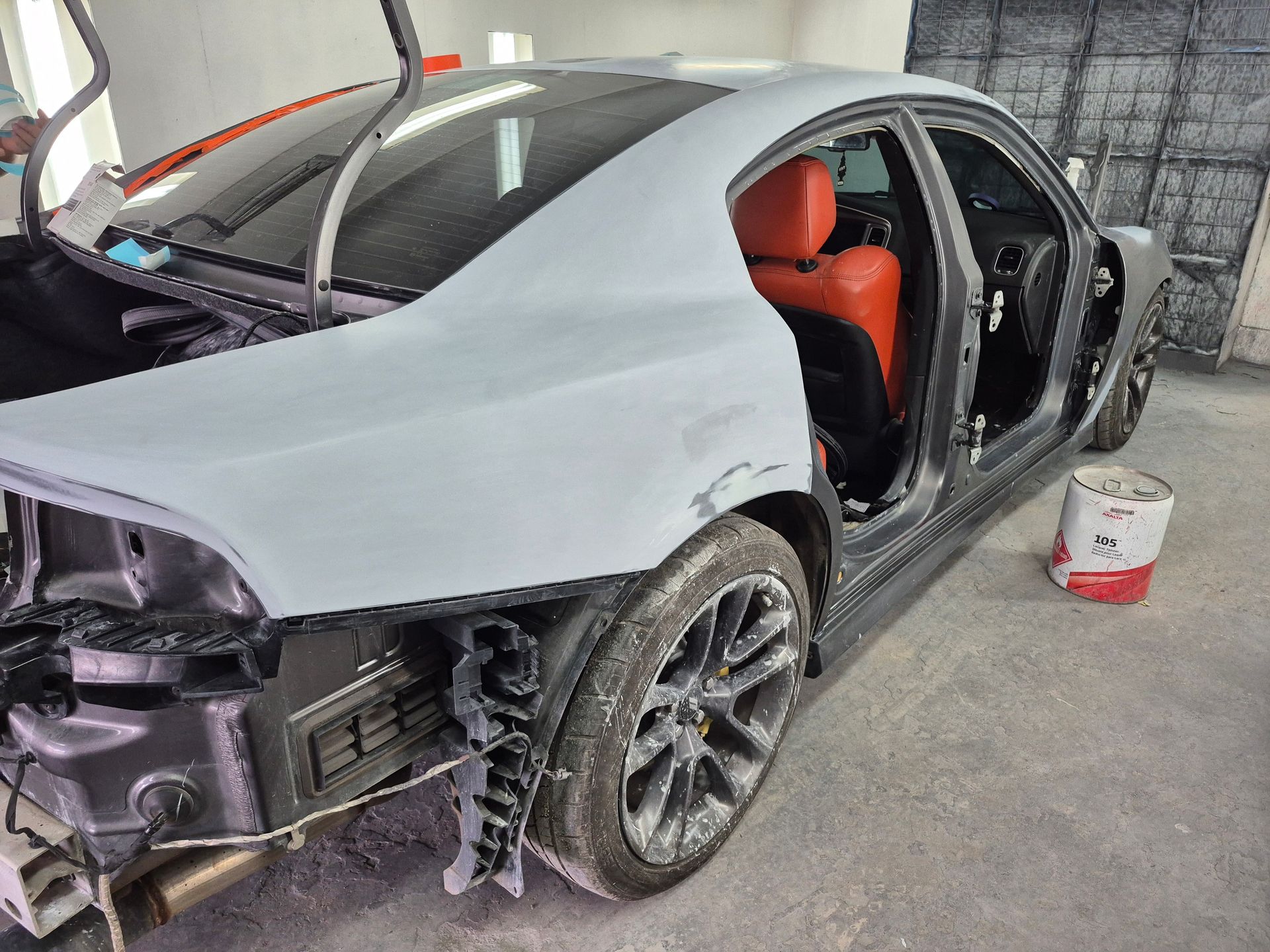 Damaged silver sports car with open doors and exposed front end in a workshop, red seats visible