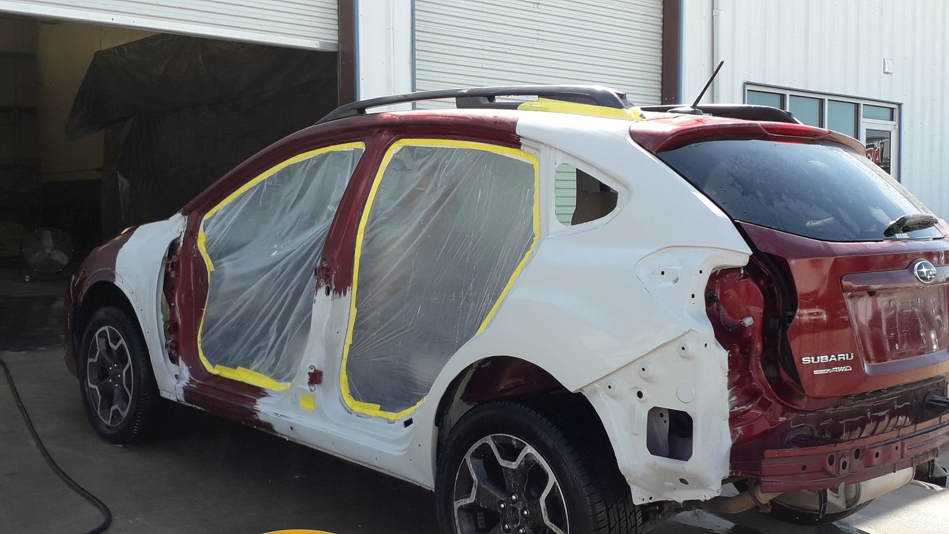 Damaged red-and-white SUV with taped plastic over windows, parked outside a garage.