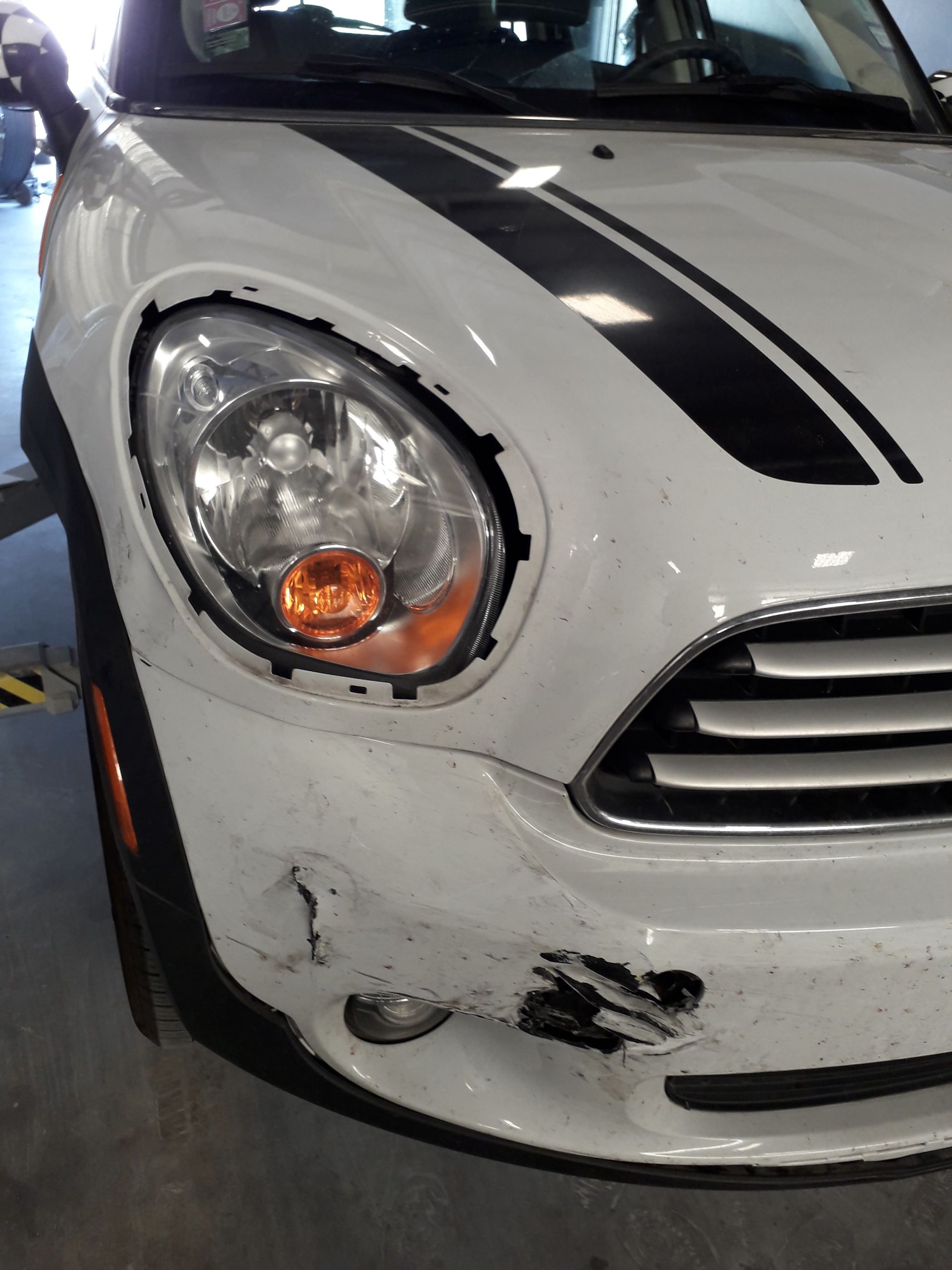 Close-up of a white car’s damaged front left corner with a cracked bumper and headlight.