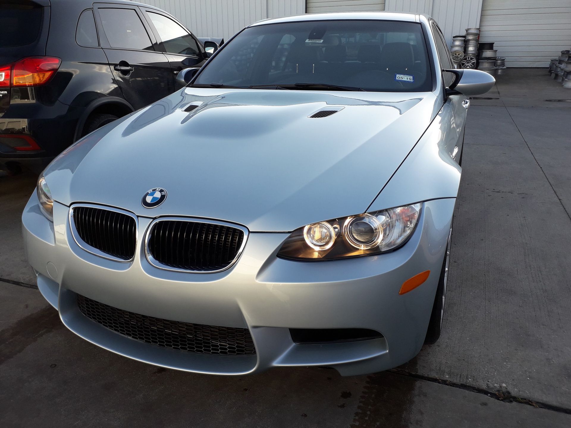 Silver BMW coupe parked outdoors on a concrete lot, front view with headlights on
