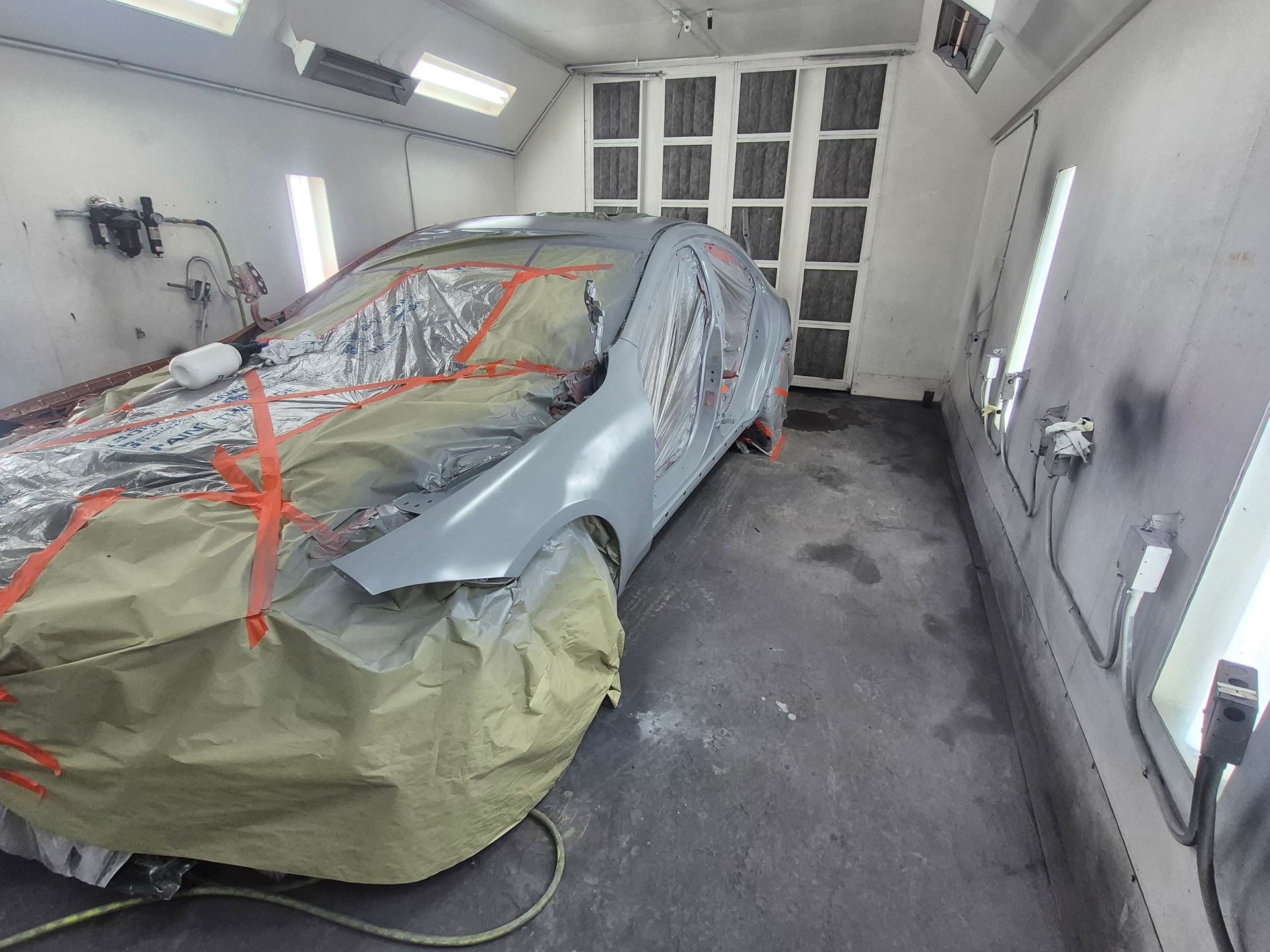 Car covered in red tape and masking paper inside a white automotive spray booth