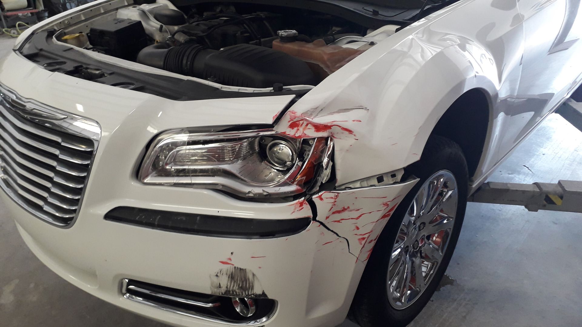 White car with front-end damage and scratches, parked indoors with the hood open