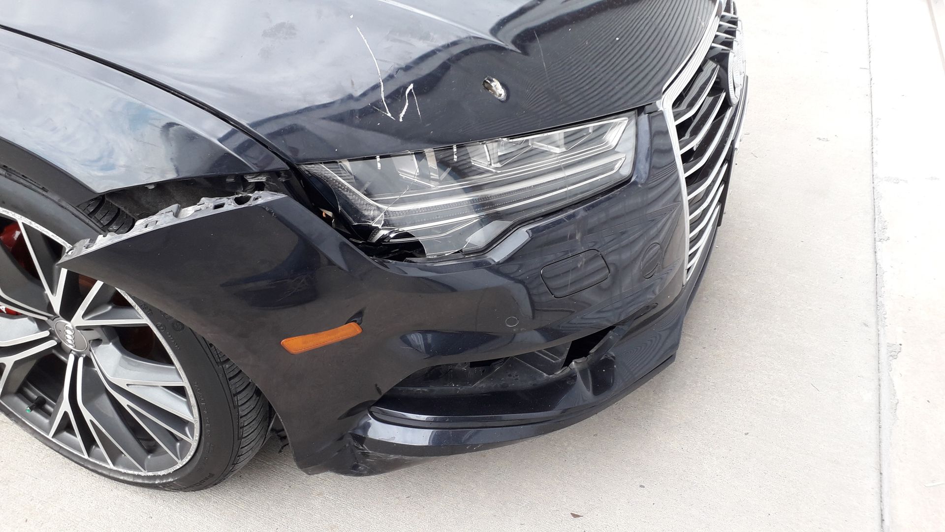 Black sedan with a damaged front bumper and fender, parked on a light-colored surface