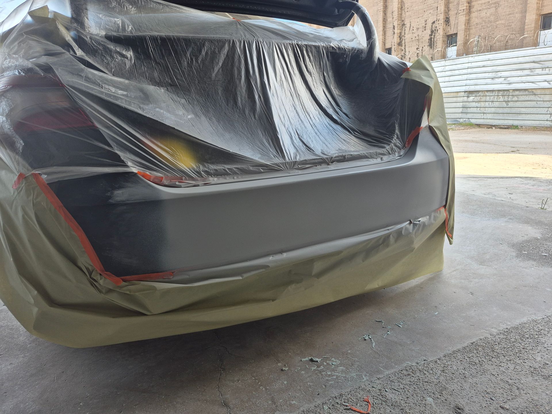 Rear of a black car masked with plastic and tape, showing bodywork repair in a garage