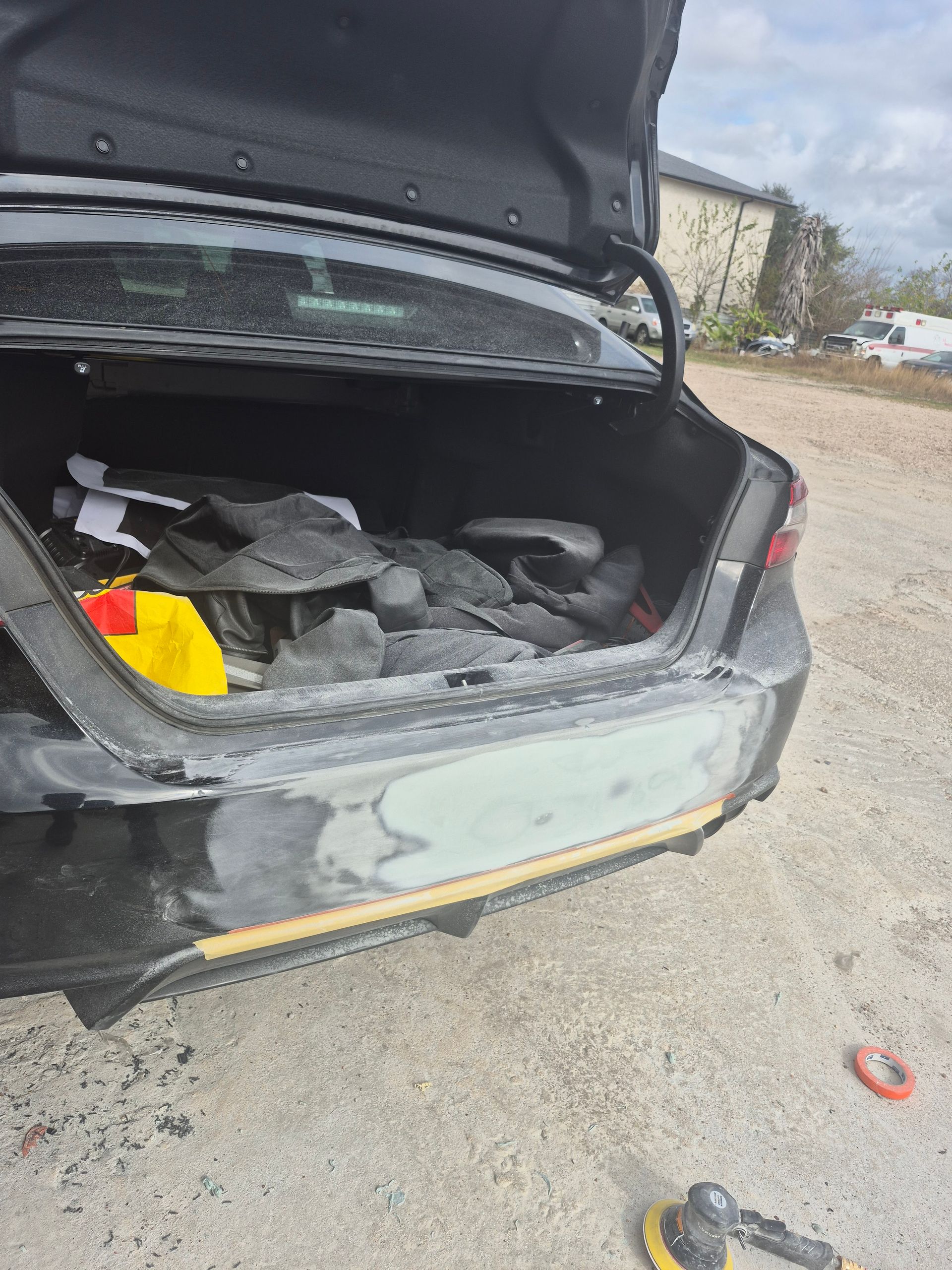 Open car trunk with damaged rear end and debris inside, parked on a gravel roadside.