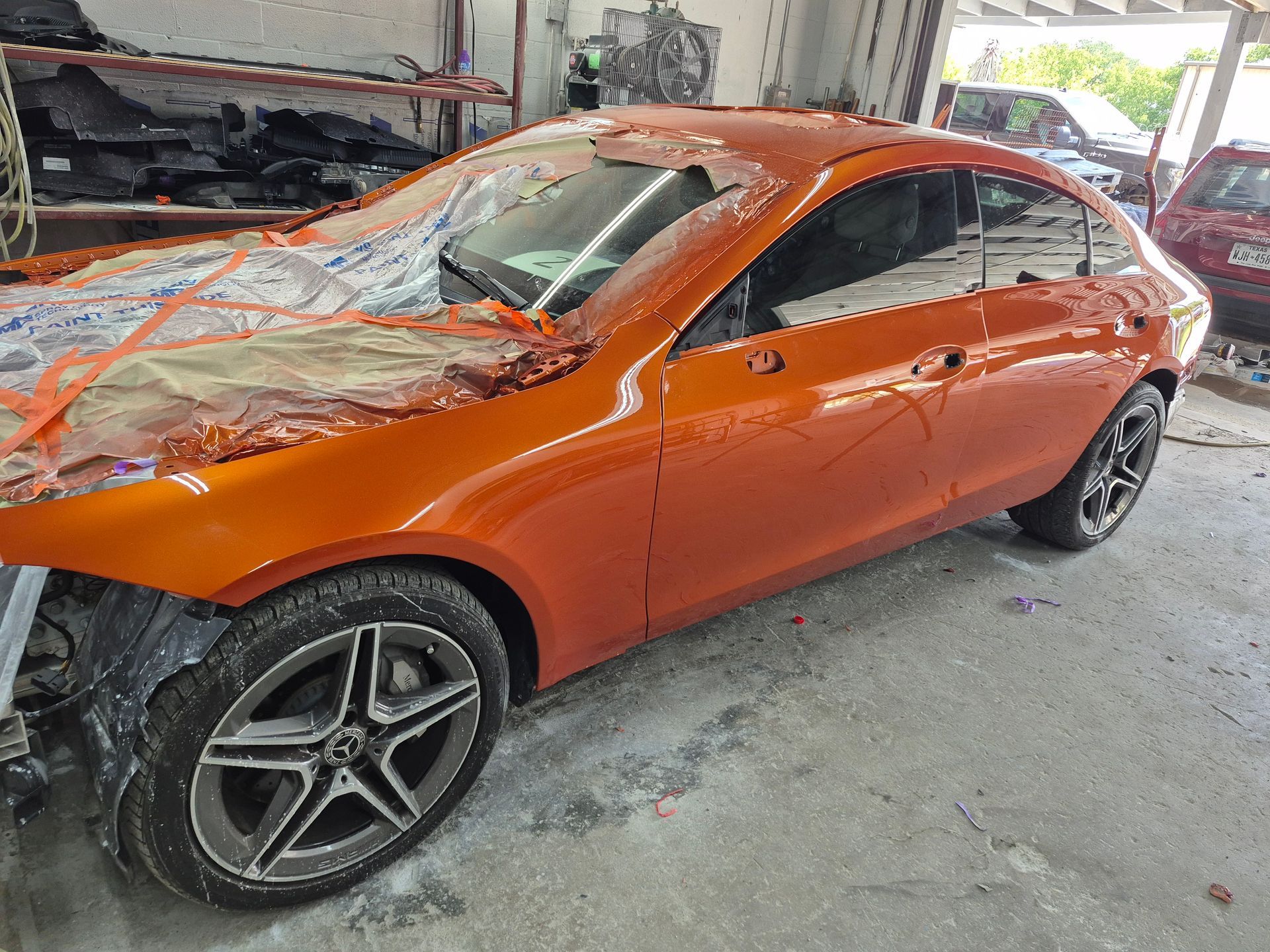 Orange sports coupe with front-end damage inside a workshop, covered in protective plastic and masking tape