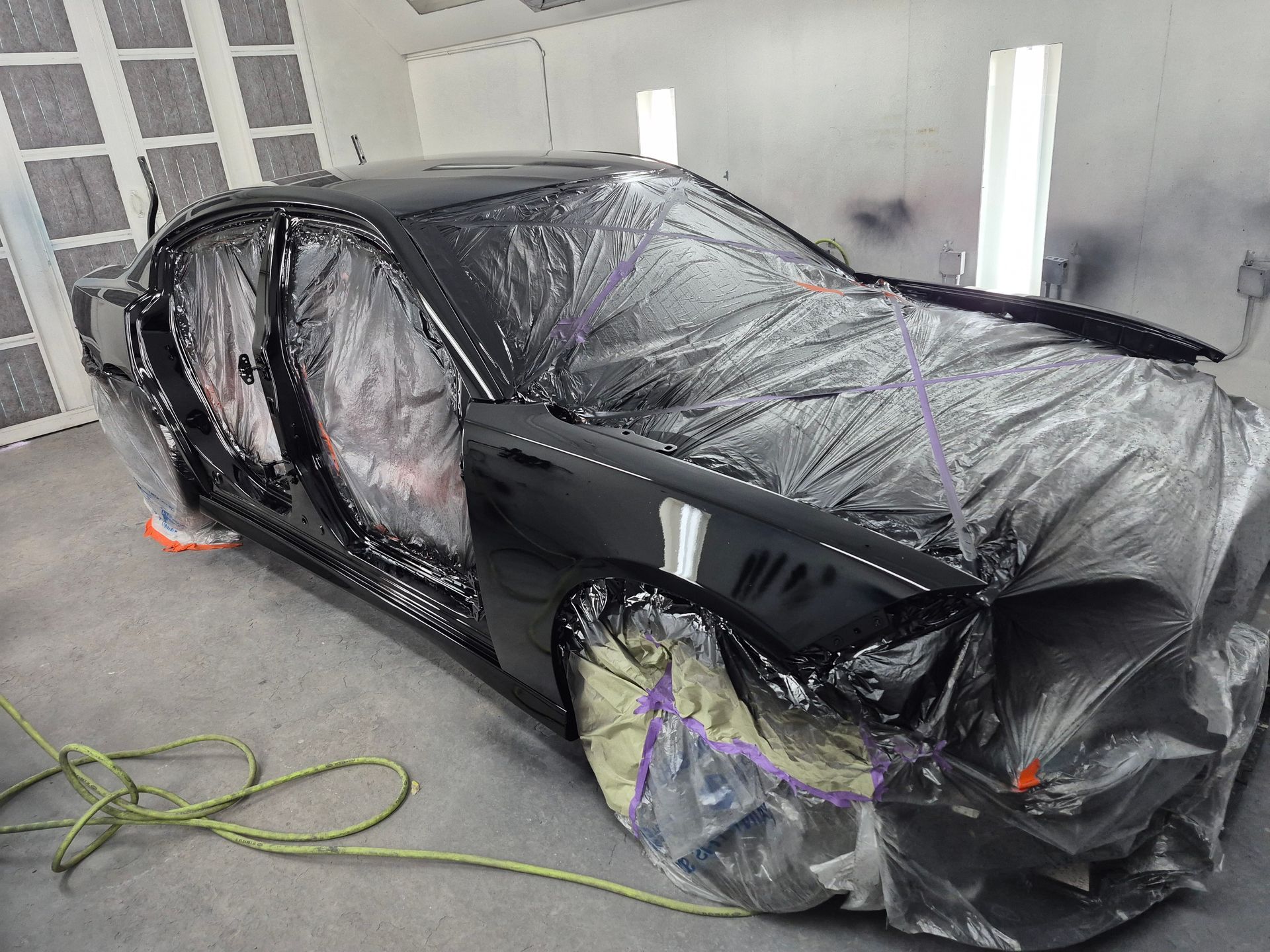 Black car body wrapped in plastic and tape in a paint booth, with a hose on the floor