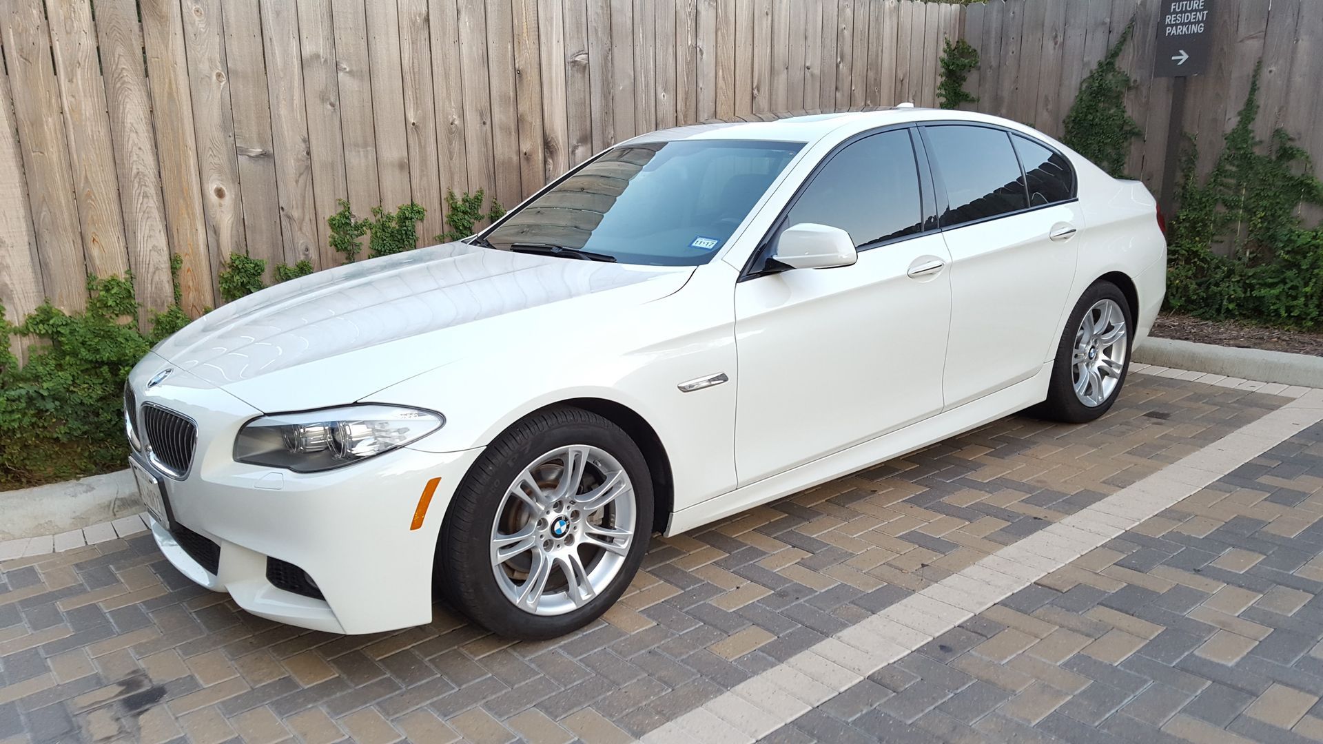 White BMW sedan parked in a driveway beside a wooden fence and brick pavers