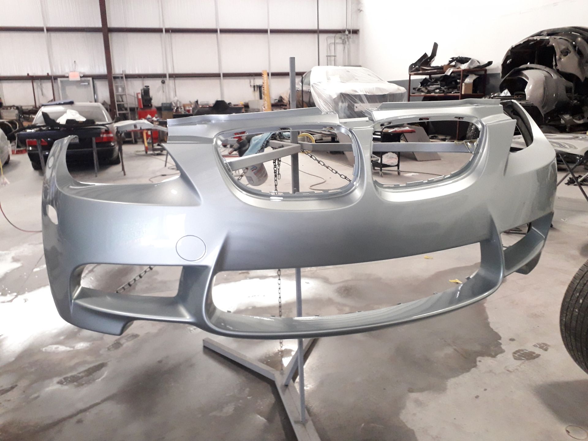 Silver car front bumper shell on a stand in a workshop, with other vehicle parts in the background