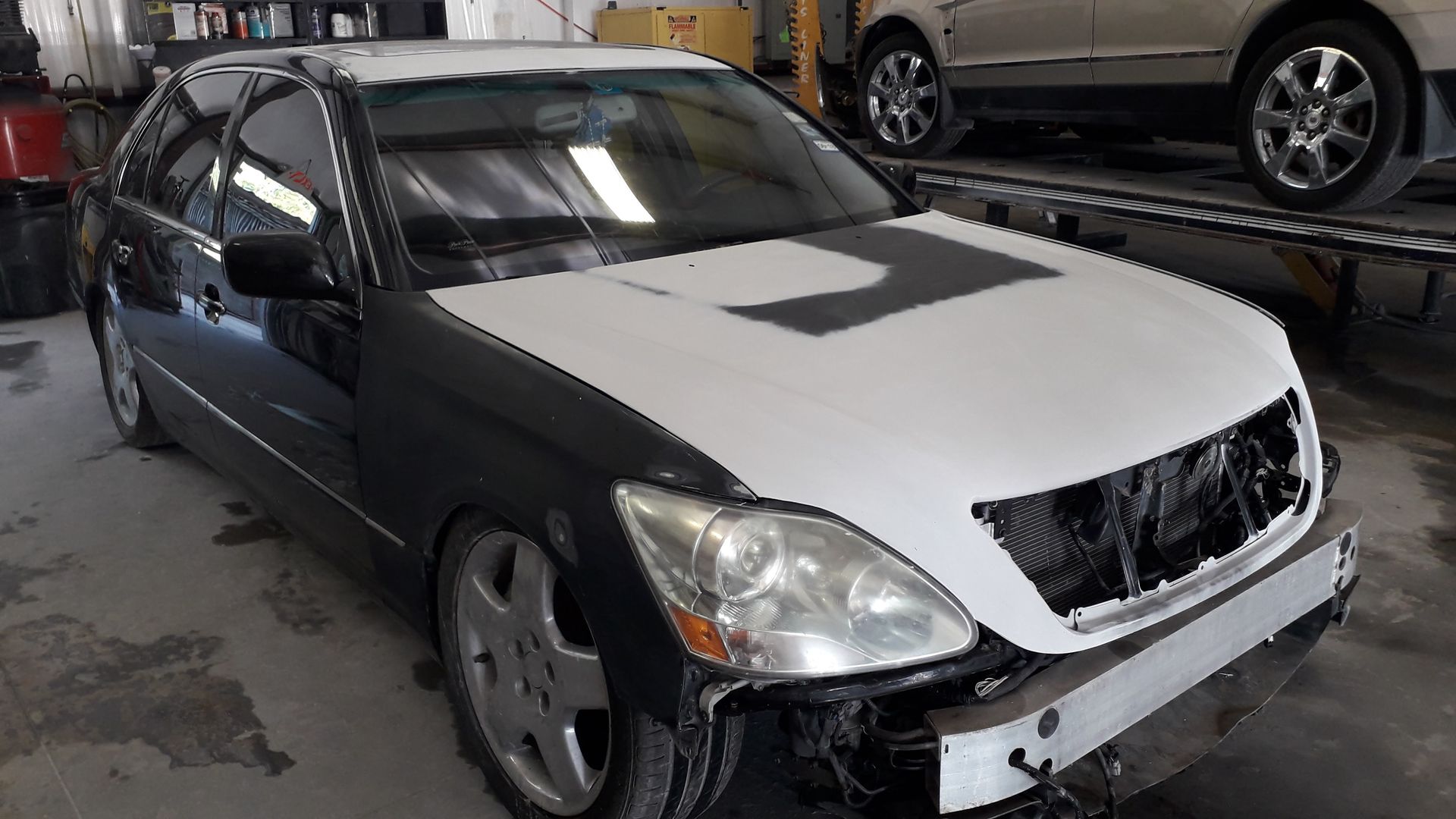 Damaged black sedan with a white hood, parked in a garage beside another car.